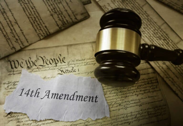 A judge’s gavel rests on old documents, including part of the U.S. Constitution. A torn piece of paper in the foreground reads 14th Amendment, symbolizing a primer on birthright citizenship.