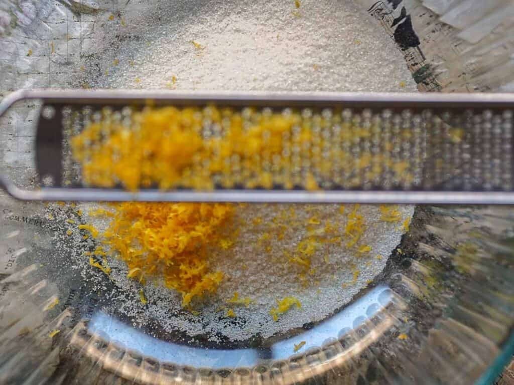 Delicious and Easy Vegan Lemon Poppyseed Muffins. 5 A close-up of a glass bowl with sugar and fresh lemon zest, with a metal zester resting across the bowl—perfect ingredients for vegan lemon poppyseed muffins.