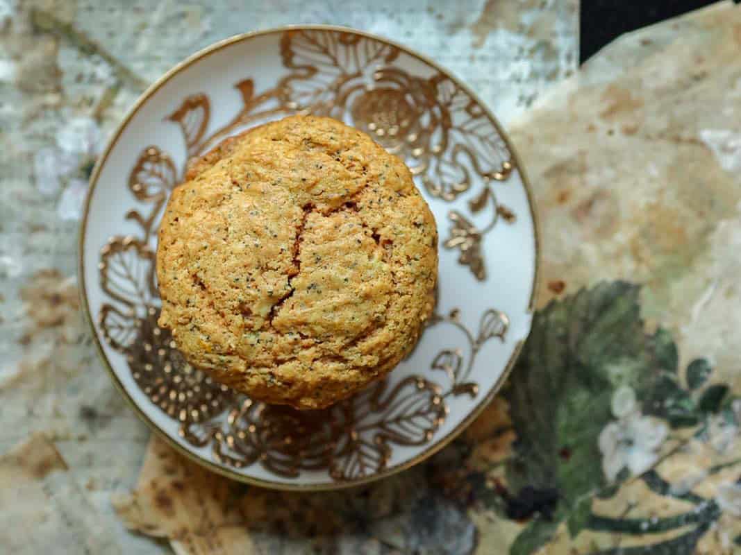 Delicious and Easy Vegan Lemon Poppyseed Muffins. 2 A golden brown, round lemon poppyseed muffin on an ornate white and gold plate, placed on a rustic, patterned surface with earthy tones and floral designs—a scene reminiscent of enjoying freshly baked vegan lemon poppyseed muffins.