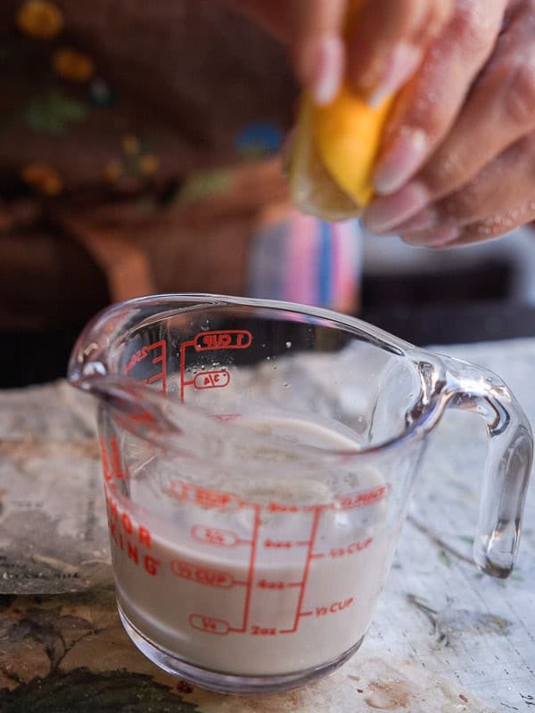 Delicious and Easy Vegan Lemon Poppyseed Muffins. 7 A hand squeezes a lemon half above a glass measuring cup of cloudy liquid, likely lemon juice mixing with milk or cream—perfect for preparing vegan lemon poppyseed muffins on a kitchen counter.