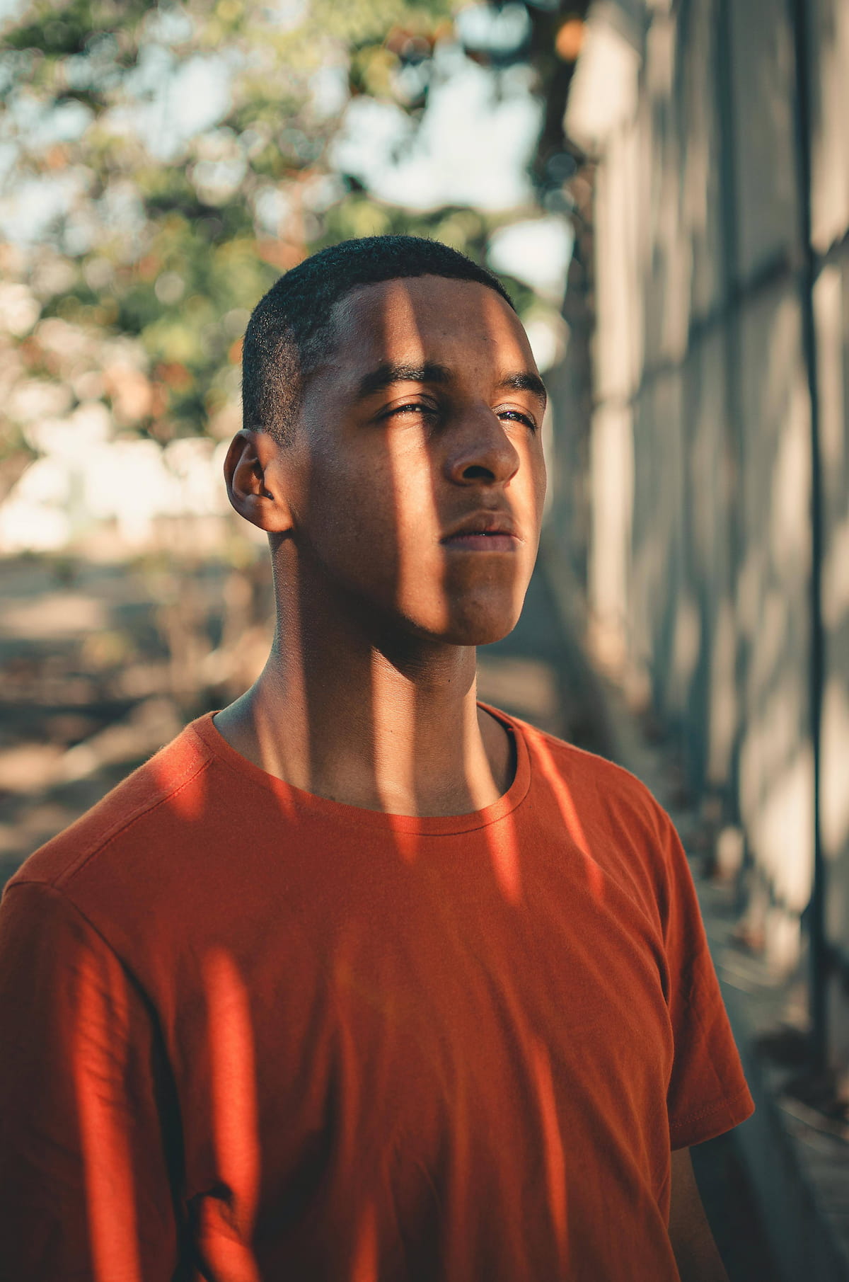 A young man with dark skin and short hair, wearing an orange t-shirt, looks contemplatively upwards and to the right. Vertical stripes of sunlight and shadow fall across his face and body.