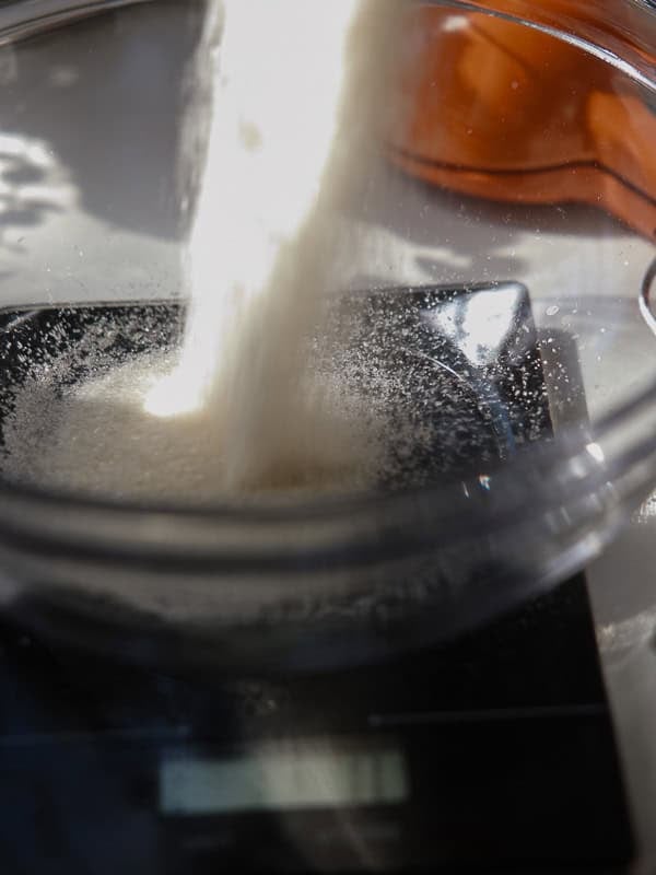 Easy Vegan Lemon Cookies Recipe (Soft and Lemony!) 5 A close-up of sugar being poured into a glass bowl on a digital kitchen scale, with sunlight illuminating the motion—perfect for making lemon cookies. A blurred orange object sits in the background.