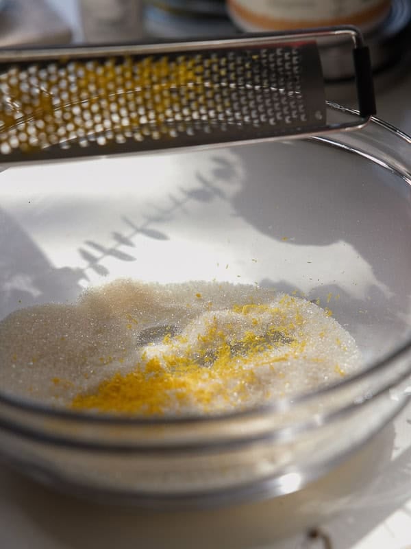 Easy Vegan Lemon Cookies Recipe (Soft and Lemony!) 7 A metal grater is held above a glass bowl filled with granulated sugar and fresh lemon zest, ready for making lemon cookies. Sunlight casts shadows and highlights the vibrant texture of the ingredients.