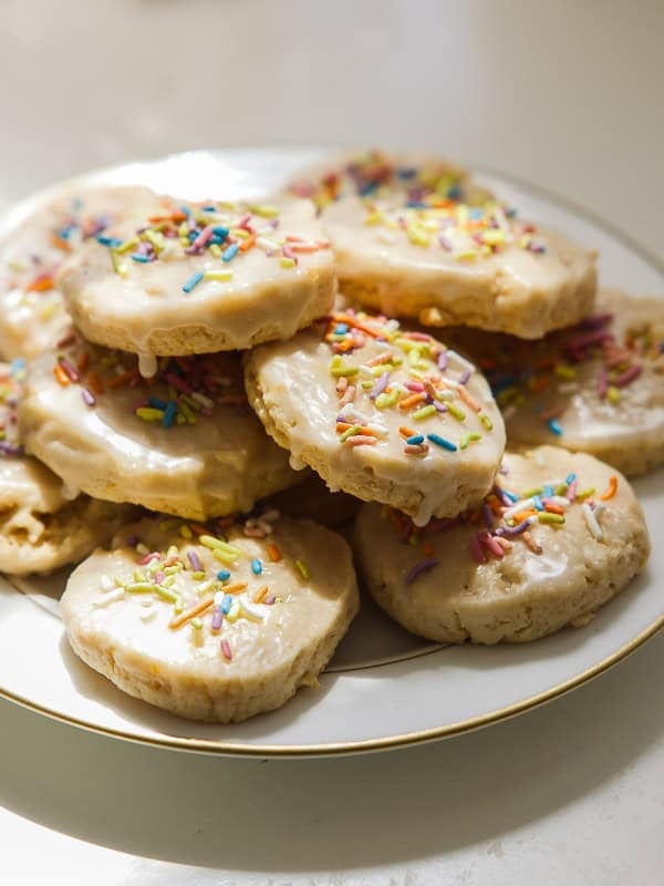 A plate of round, glazed lemon cookies topped with colorful sprinkles, stacked in a pile on a white surface with sunlight shining on them.