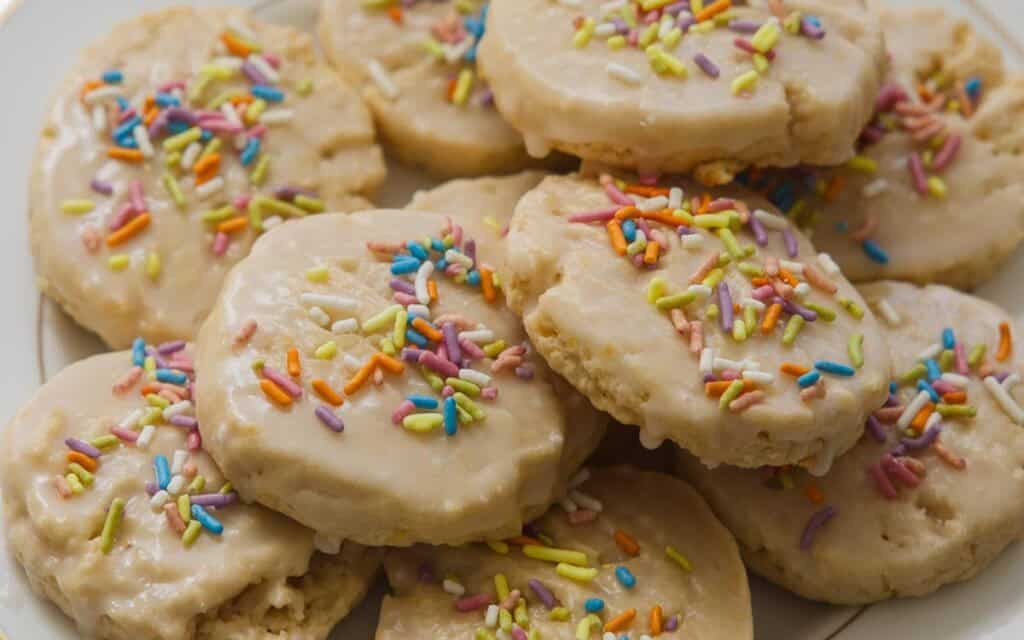 A plate of frosted lemon cookies topped with colorful rainbow sprinkles, arranged in a pile. The cookies have a soft texture and a glossy layer of icing.