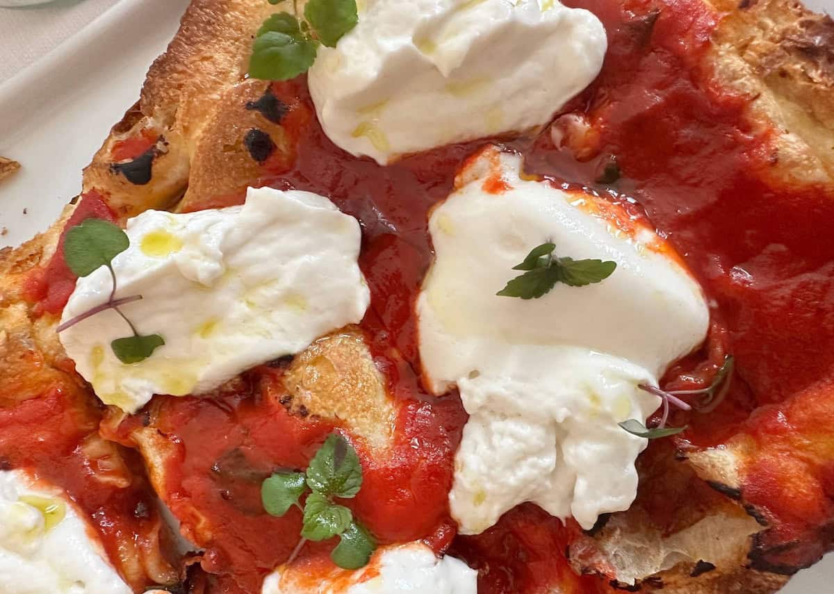 Da Vinci & Mutti: A Visit to Parma, Italy. 2 Close-up of a rustic pizza topped with creamy burrata cheese, vibrant Mutti tomato sauce, microgreens, and olive oil on a golden, slightly charred crust—reminiscent of flavors from Parma, Italy.