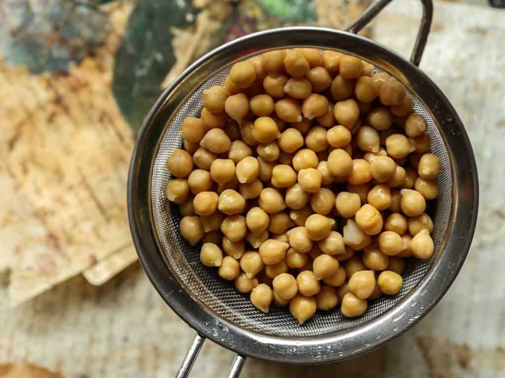 A metal strainer filled with rinsed chickpeas, perfect for making chickpea cutlets, sits over a bowl on a rustic surface.