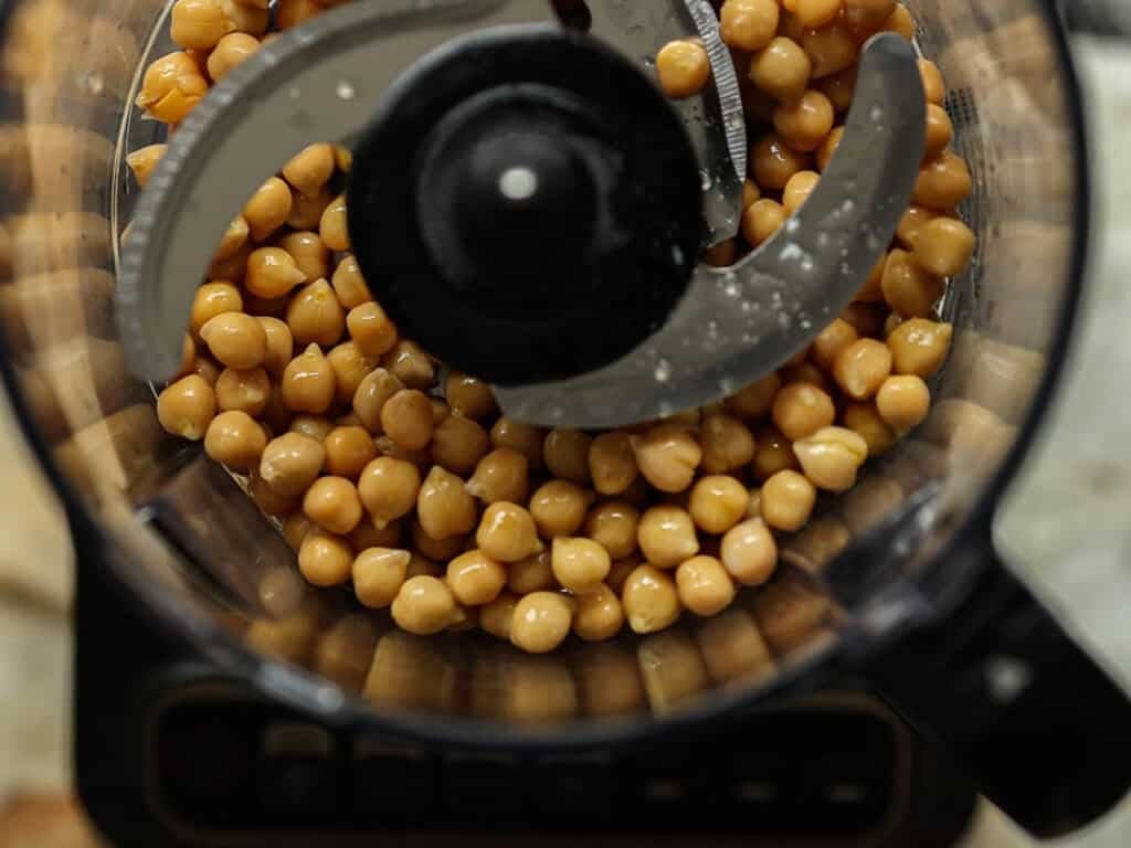 Overhead view of chickpeas inside a food processor with metal blades, ready to be blended for homemade chickpea cutlets.