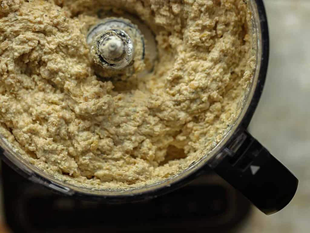 Close-up view of a food processor filled with a thick, beige mixture for chickpea cutlets, with textured swirls around the central blade, suggesting a well-blended batter or dough.