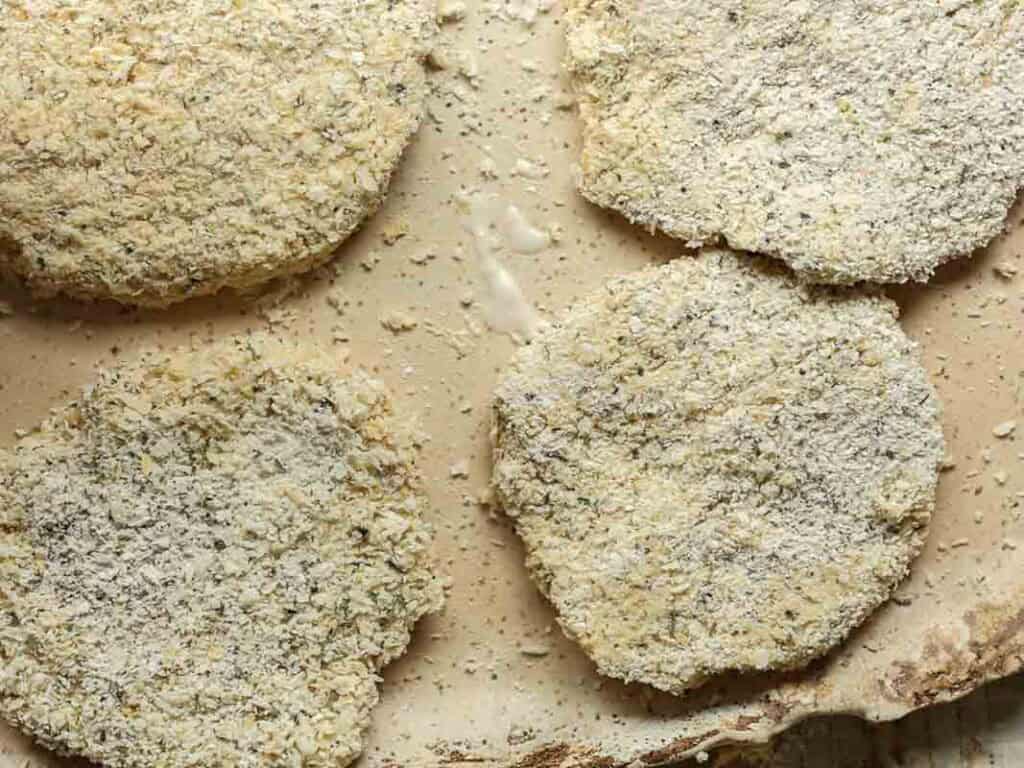 Four breaded, uncooked chickpea cutlets are arranged on a light-colored surface, with some crumbs scattered around them. The patties appear to be coated in seasoned breadcrumbs, ready for baking or frying.