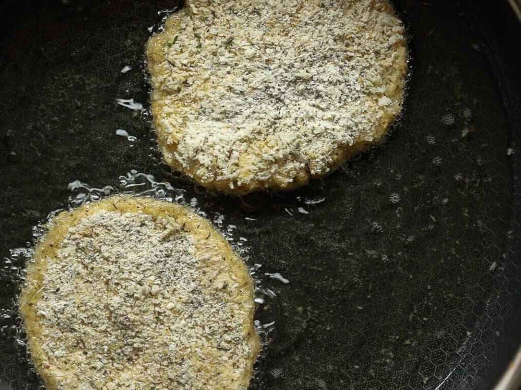 Two breaded chickpea cutlets are frying in oil in a dark nonstick pan, with visible bubbles forming around the edges of the patties.