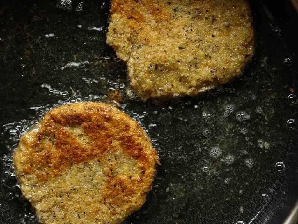 Two golden-brown chickpea cutlets are frying in oil in a dark pan, with visible bubbles around the edges, indicating they are being cooked.