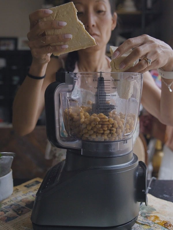 A woman adds chunks of tofu to a food processor already filled with chickpeas, preparing homemade chickpea cutlets in a kitchen setting.