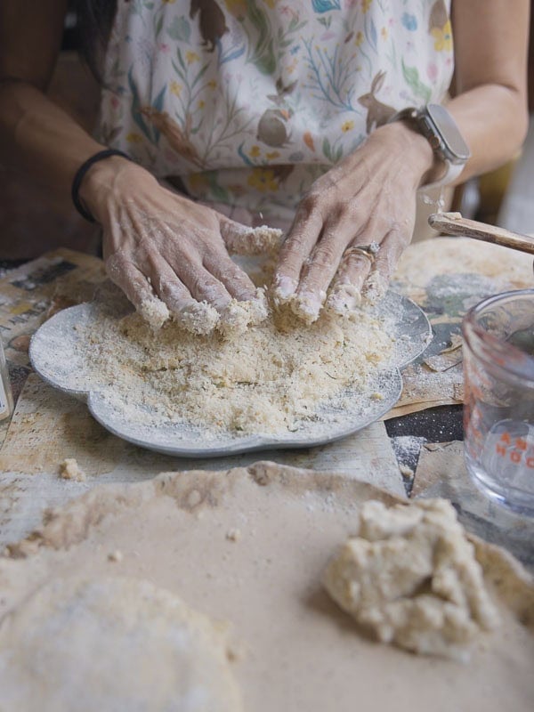 A person wearing a patterned apron mixes flour and dough for chickpea cutlets with their hands on a plate, surrounded by baking tools and ingredients on a kitchen counter.