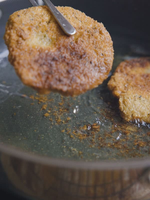 A chickpea cutlet is being lifted from a frying pan with oil using a metal utensil, while another cutlet remains in the pan. The chickpea cutlets are golden brown and crispy.