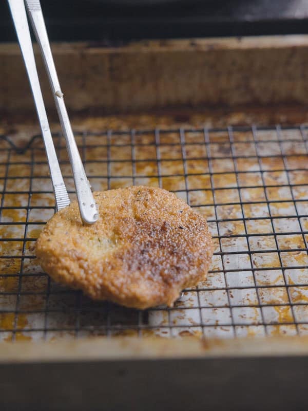A pair of metal tongs holds a golden-brown, breaded chickpea cutlet on a wire rack set over a baking tray, likely cooling after frying.