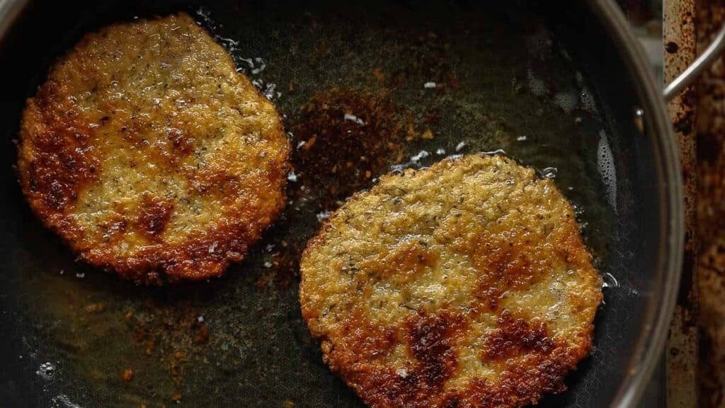Two golden-brown chickpea cutlets frying in oil in a dark skillet, with visible crispy edges and a textured surface.