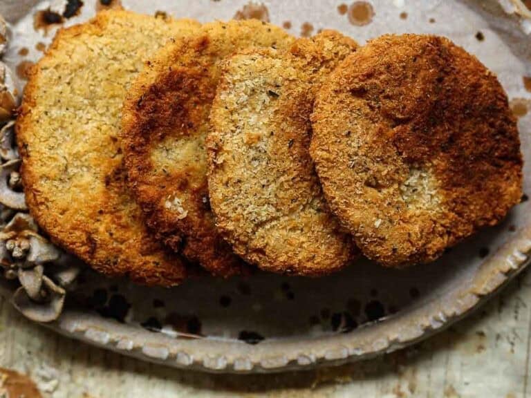 Four golden-brown, crispy chickpea cutlets are arranged in a row on an oval ceramic plate, viewed from above, with rustic textures and light brown spots on the plate.