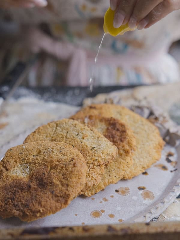 A hand squeezes a lemon over three pieces of breaded, fried chickpea cutlets on a round tray, with crumbs scattered around. The background is slightly blurred, showing a person in patterned clothing.