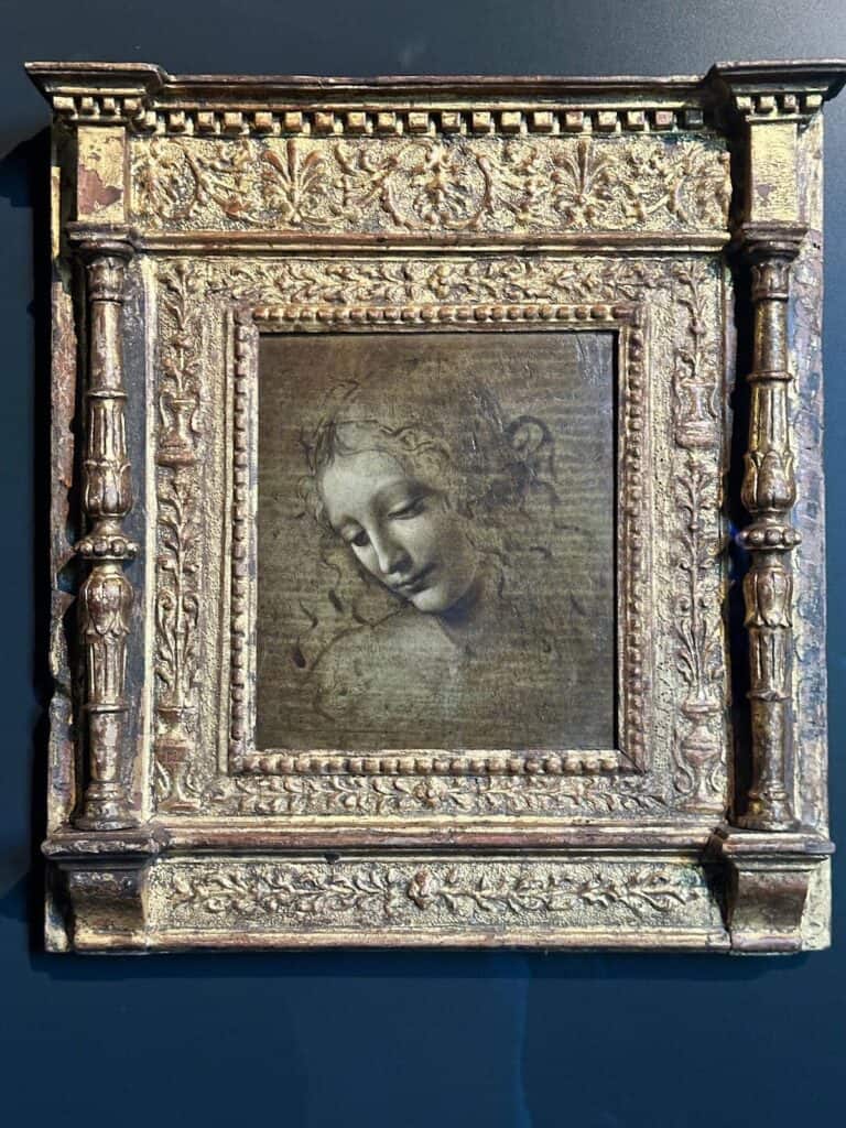 Da Vinci & Mutti: A Visit to Parma, Italy. 8 A framed artwork featuring a delicate drawing of a young woman's face, looking downward and slightly smiling, in an ornate, gold, carved Renaissance-style frame against a dark background—evoking the timeless elegance of Da Vinci in Parma, Italy.