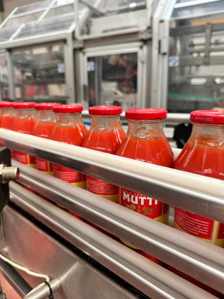 Da Vinci & Mutti: A Visit to Parma, Italy. 16 Glass bottles of Mutti tomato sauce with red caps are aligned on a conveyor belt inside a factory in Parma, with industrial equipment visible in the background.