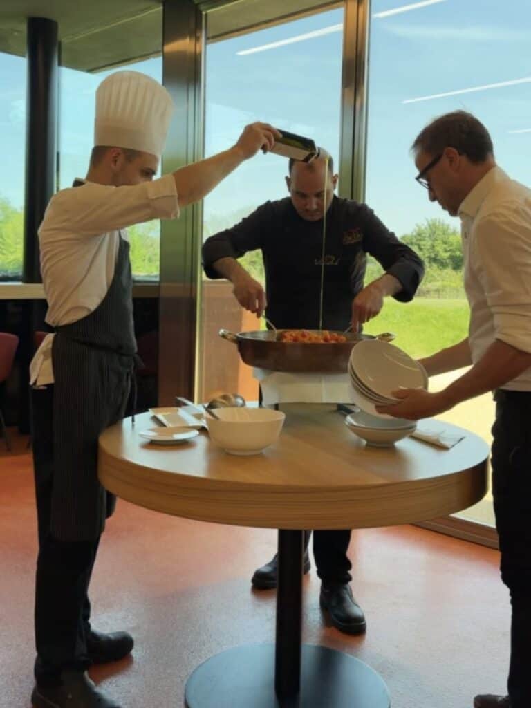 Da Vinci & Mutti: A Visit to Parma, Italy. 23 Three chefs stand around a round table indoors. One pours Mutti oil, another mixes food in a large pan, and the third holds several stacked white bowls. Bright daylight streams in through large windows behind them.