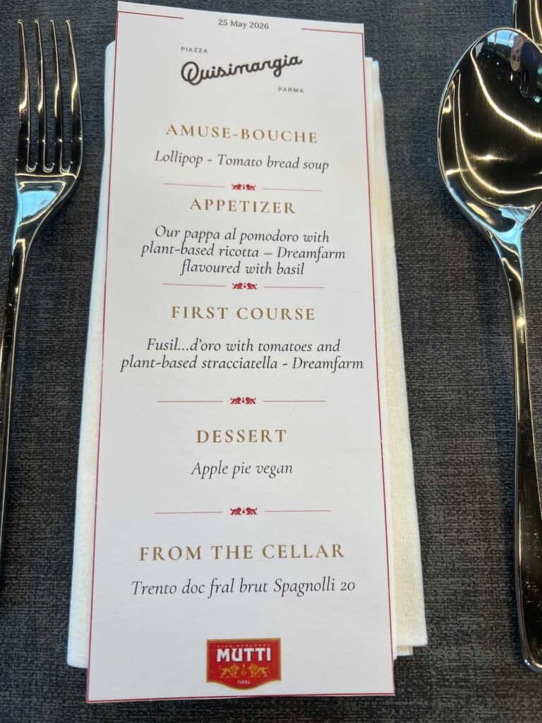 Da Vinci & Mutti: A Visit to Parma, Italy. 24 A restaurant menu on a white napkin, featuring dishes like amuse-bouche (tomato bread soup lollipop), appetizer (Mutti tomato soup with plant-based ricotta and basil), fusilli with vegan stracciatella, vegan apple pie, and sparkling wine.