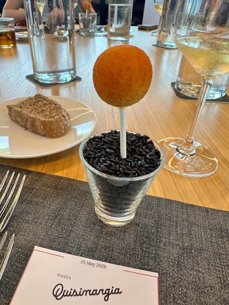Da Vinci & Mutti: A Visit to Parma, Italy. 19 A round, Parma-breaded appetizer on a stick is served upright in a cup filled with black rice, next to a slice of bread, glasses of wine, and a menu on a wooden table.
