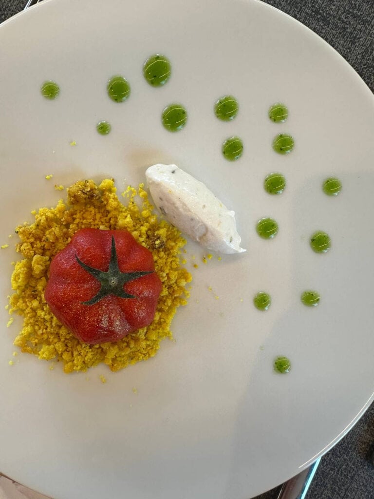 Da Vinci & Mutti: A Visit to Parma, Italy. 20 A white plate features a red, Mutti tomato-shaped item on yellow crumbs, a quenelle of cream, and green sauce dots arranged artistically on the right side.