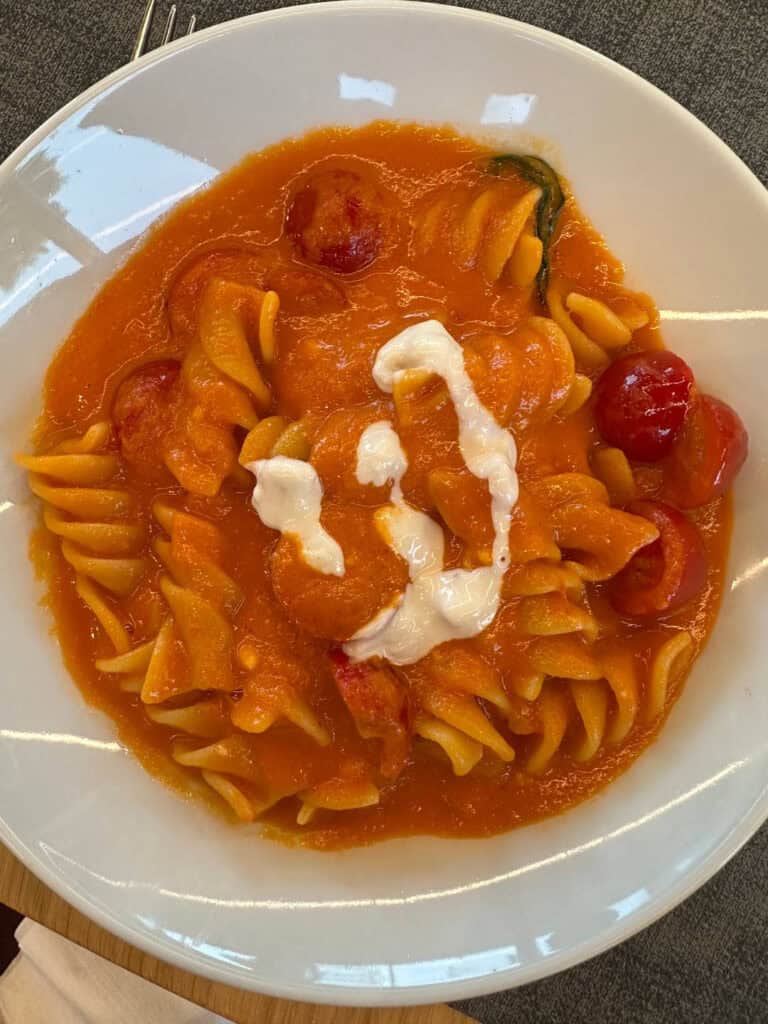 Da Vinci & Mutti: A Visit to Parma, Italy. 21 A white plate of fusilli pasta in tomato sauce, topped with a swirl of melted cheese and Parma cherry tomatoes, set on a gray tablecloth—bringing the rich flavors of Italy to your table.