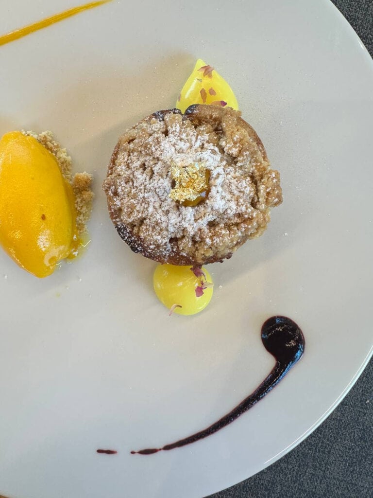 Da Vinci & Mutti: A Visit to Parma, Italy. 22 A plated dessert inspired by Da Vinci, featuring a crumb-topped pastry dusted with powdered sugar, a scoop of bright yellow sorbet, yellow cream dollops, a swirl of dark sauce, and caramel drizzle on a white plate.