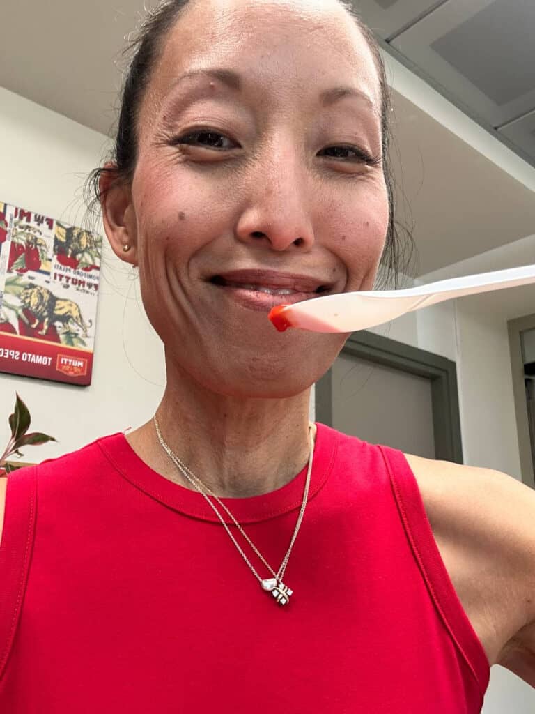 Da Vinci & Mutti: A Visit to Parma, Italy. 18 A woman wearing a red sleeveless top and necklace smiles while holding a plastic spoon with Mutti red sauce near her mouth. She stands indoors with a plant and posters visible in the background.