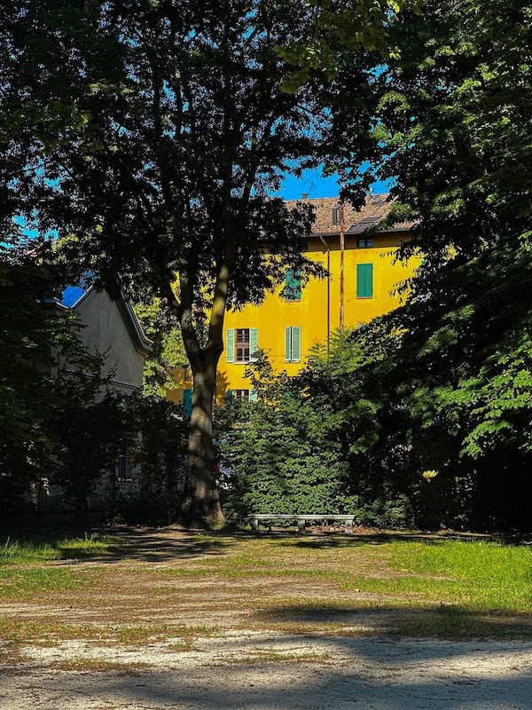 Da Vinci & Mutti: A Visit to Parma, Italy. 4 A bright yellow building with green shutters, reminiscent of Parma Italy, is partially obscured by tall trees casting shadows on the ground in a sunlit park setting.