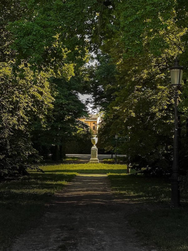 Da Vinci & Mutti: A Visit to Parma, Italy. 3 A tree-lined path in Parma, Italy, leads to a stone urn monument bathed in sunlight, with a building visible in the background and a vintage lamp post on the right side, evoking a scene Da Vinci himself might have admired.