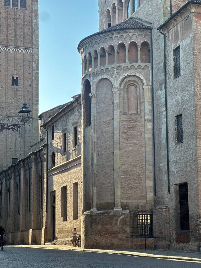 A sunlit street in Parma, Italy beside an old church with tall brick walls, arched windows, and decorative columns. A person stands near a bicycle by the building.
