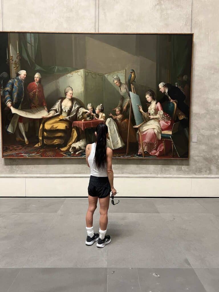 A person with long dark hair in a white tank top and black shorts stands in a museum, looking at a large classical painting by Mutti depicting several elegantly dressed people gathered indoors.