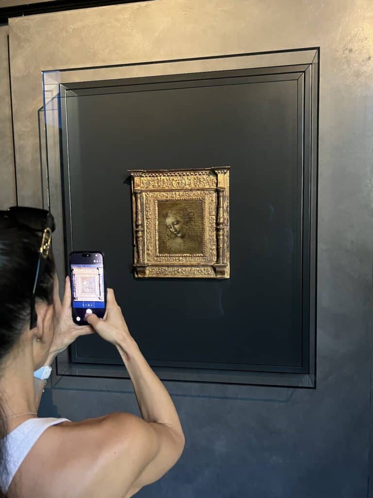A person takes a photo with a smartphone of a small, framed portrait—possibly of their Mutti—displayed on a dark wall in a gallery. The sepia-toned drawing is set in an ornate gold frame, capturing both art and sentiment.