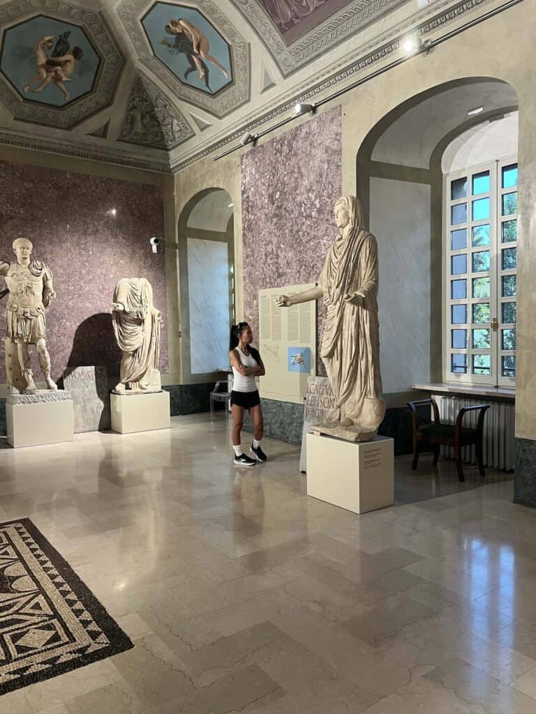 A woman stands in a museum room observing a marble statue among other classical sculptures, with tall windows and detailed ceiling artwork in the background—her thoughtful gaze reminiscent of Mutti's quiet admiration for art.