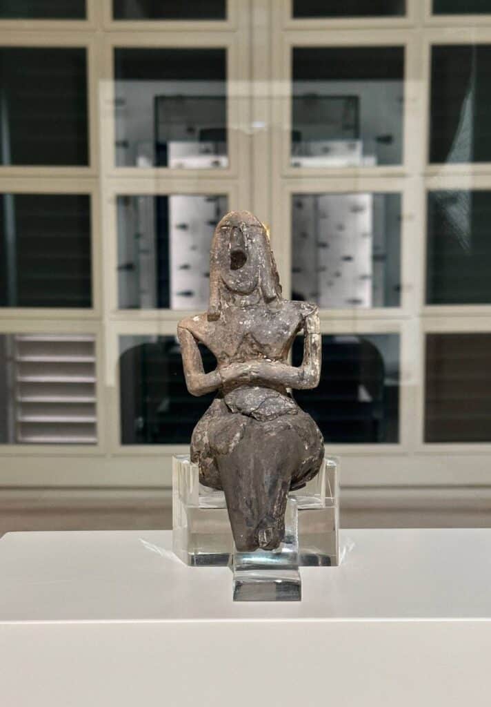 Da Vinci & Mutti: A Visit to Parma, Italy. 7 A stone figurine of a seated woman with pronounced hips and crossed arms is displayed on a clear stand in a museum setting.