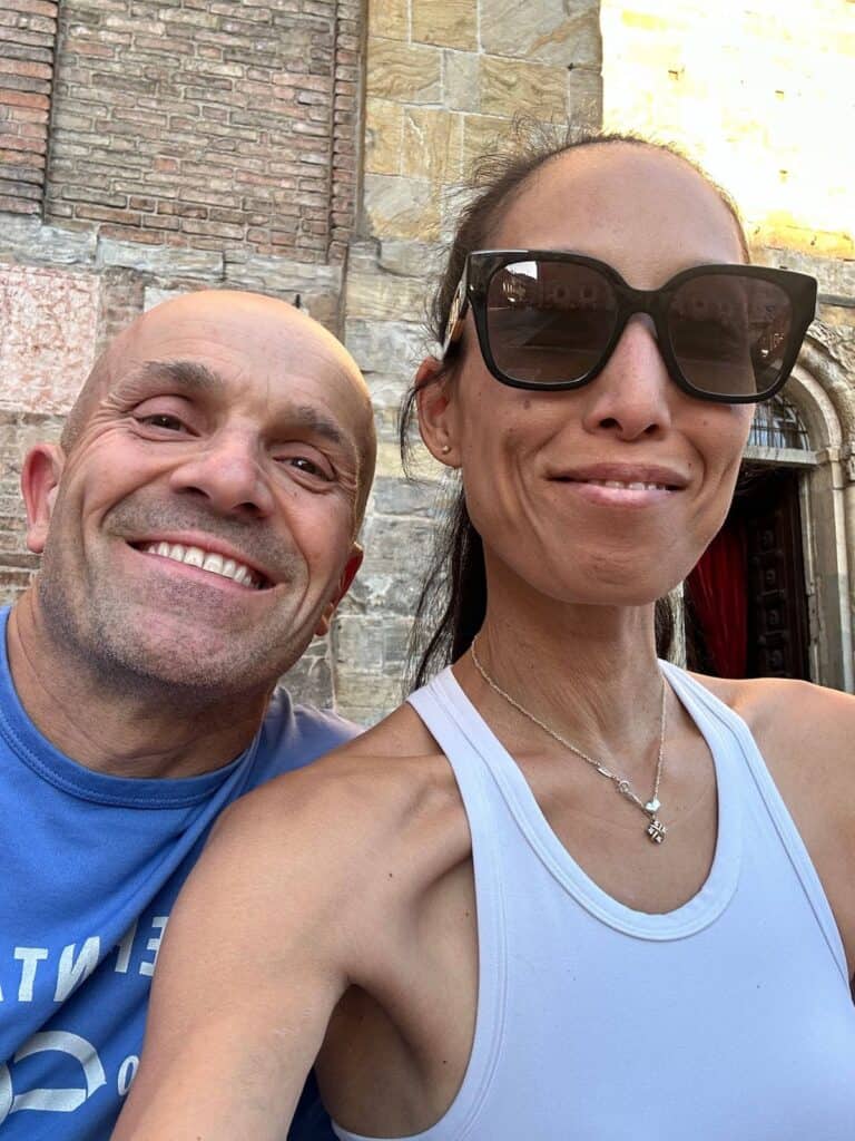 A man and a woman smile for a selfie outdoors in front of an ornate stone building. The man wears a blue shirt, while Mutti sports sunglasses and a white top. Both appear happy and relaxed.