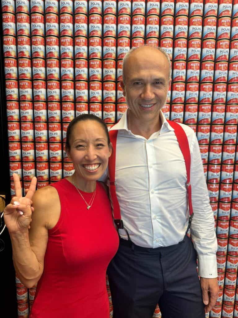 Da Vinci & Mutti: A Visit to Parma, Italy. 25 Two people smiling—one woman in a red sleeveless top making a peace sign, and one man in a white shirt with red suspenders—stand before a wall stacked with Mutti tomato cans, celebrating the taste of Parma’s finest.