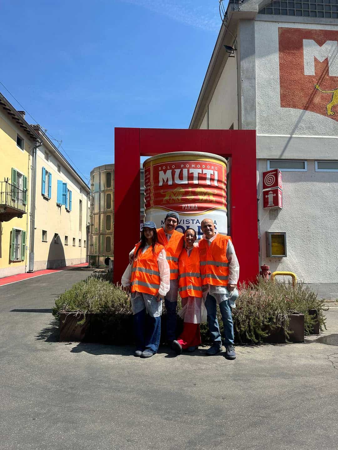 Four people wearing orange safety vests and hairnets stand smiling in front of a large Mutti tomato can display, celebrating the Parma tradition, outside a building on a sunny day.