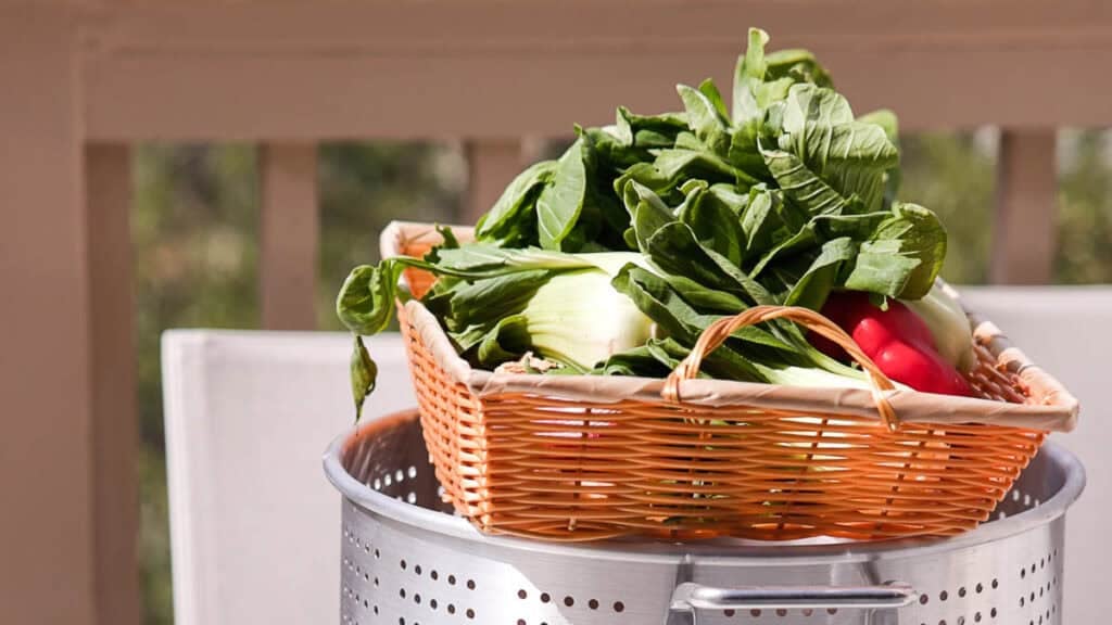BEST Baby Bok Choy Kimchi Recipe - Easy & Delicious 4 A wicker basket filled with leafy greens, radishes, and fresh bok choy kimchi sits on top of a large metal colander outdoors, with a blurred background of a porch railing.