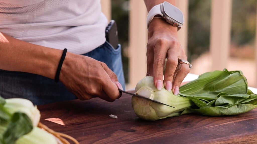 BEST Baby Bok Choy Kimchi Recipe - Easy & Delicious 17 A person wearing a white watch slices bok choy on a wooden cutting board outdoors, preparing fresh ingredients for homemade bok choy kimchi.