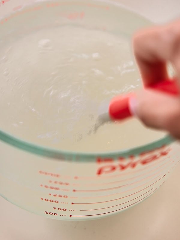 BEST Baby Bok Choy Kimchi Recipe - Easy & Delicious 23 A hand stirs a clear liquid in a large glass measuring cup marked with red measurement lines, preparing the brine for bok choy kimchi.
