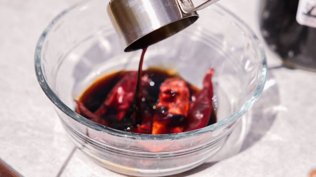 BEST Baby Bok Choy Kimchi Recipe - Easy & Delicious 22 A metal measuring cup pours dark liquid over dried red chilies in a clear glass bowl, sitting on a light-colored surfaceโan essential step when preparing flavorful bok choy kimchi.