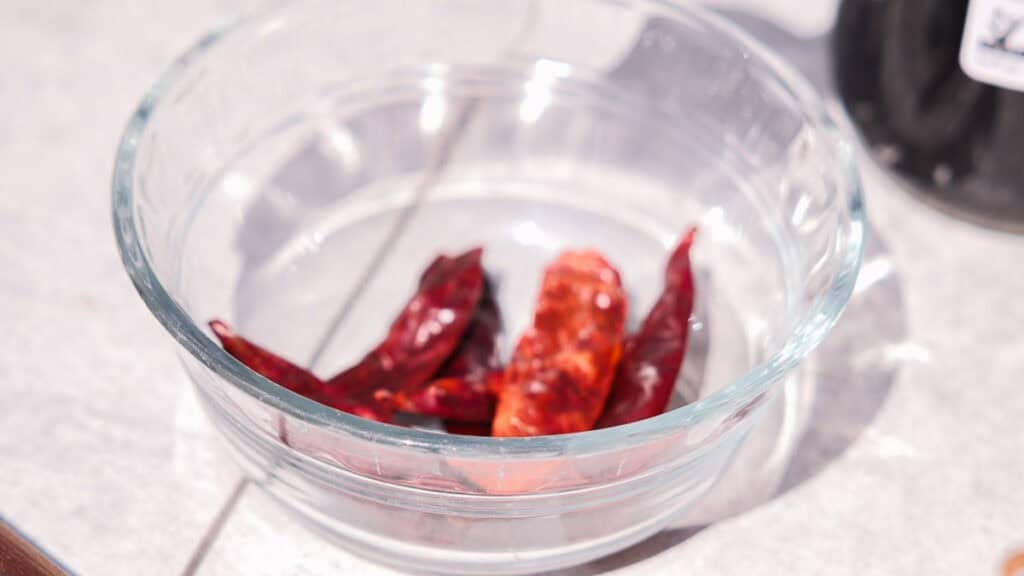 BEST Baby Bok Choy Kimchi Recipe - Easy & Delicious 16 Clear glass bowl containing several dried red chili peppers and a hint of bok choy kimchi, placed on a white countertop with sunlight casting shadows.