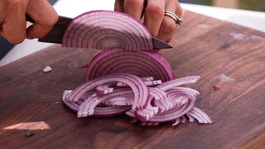 BEST Baby Bok Choy Kimchi Recipe - Easy & Delicious 8 A person slices a red onion on a wooden cutting board with a knife, creating thin, even rings to accompany a fresh batch of bok choy kimchi.