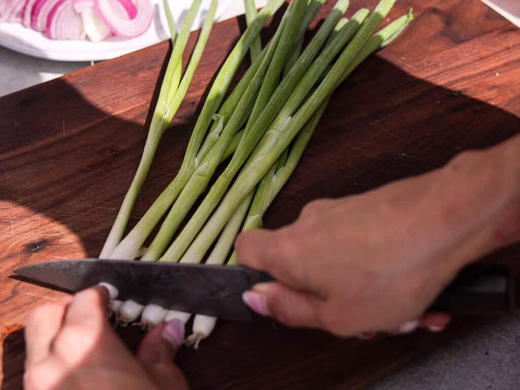 BEST Baby Bok Choy Kimchi Recipe - Easy & Delicious 9 A person slicing fresh green onions on a wooden cutting board with a knife; a plate of sliced red onions and freshly made bok choy kimchi is in the background.