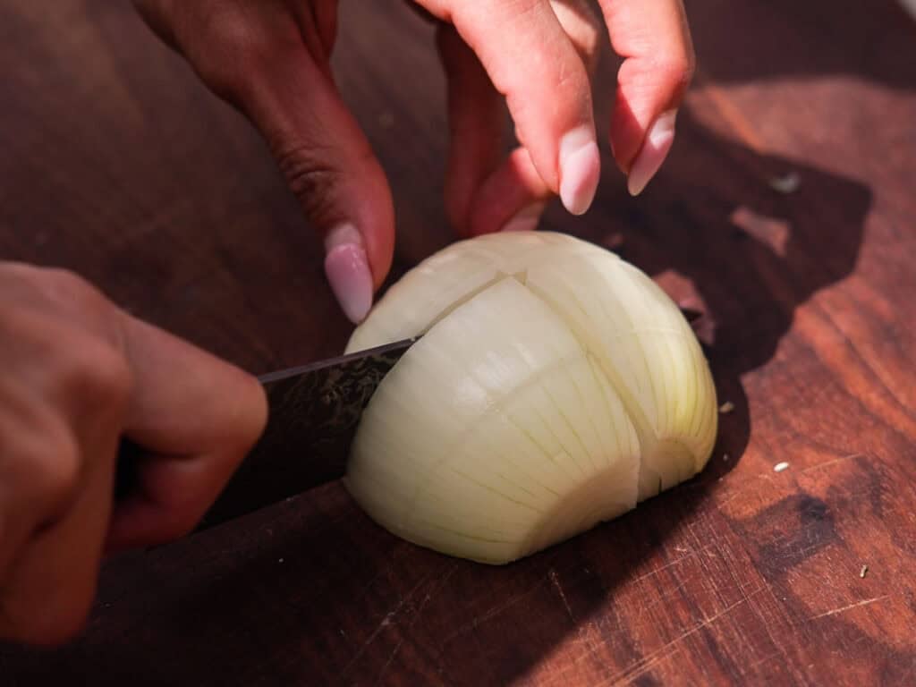 BEST Baby Bok Choy Kimchi Recipe - Easy & Delicious 7 A person slices a half onion with a knife on a wooden cutting board, using one hand to hold the onion and the other to cut, preparing ingredients for homemade bok choy kimchi.
